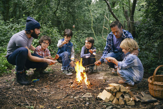Two fathers and four boys toasting marshmallows on campfire in forest — Stock Photo