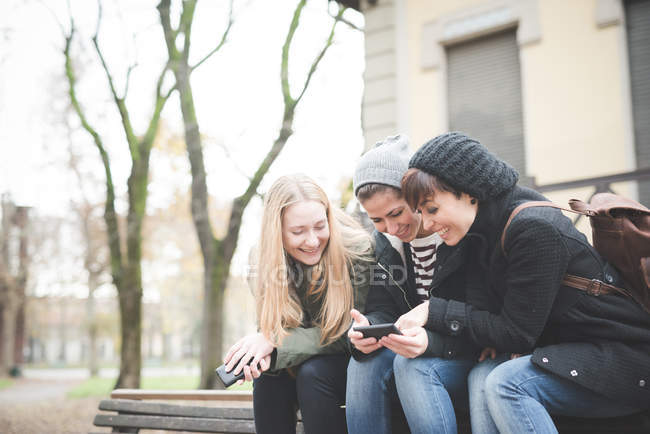 Três irmãs usando smartphone no banco do parque — Fotografia de Stock