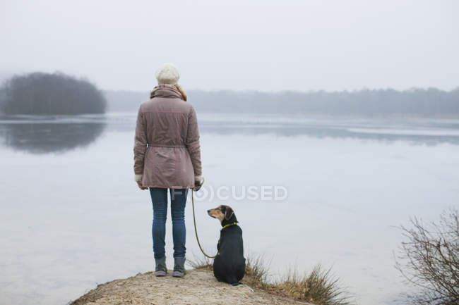 Rückansicht einer Frau mit Hund, die vom Flussufer aus blickt — Stockfoto