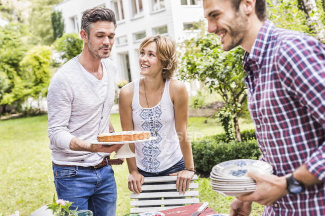 Группа друзей устраивает вечеринку в саду, средний взрослый мужчина несет торт — стоковое фото