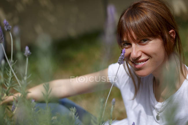 Mujer adulta sentada entre lavanda, mirando a la cámara, sonriendo - foto de stock