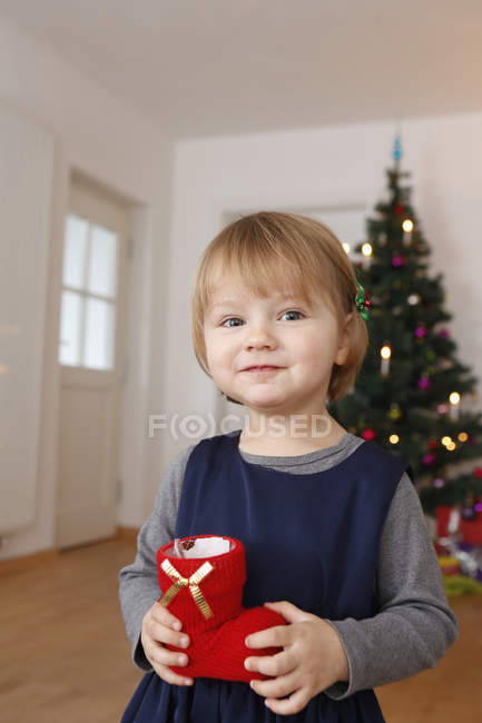 Дівчина перед різдвяною ялинкою тримає червоний черевик, дивлячись на камеру посміхаючись — стокове фото