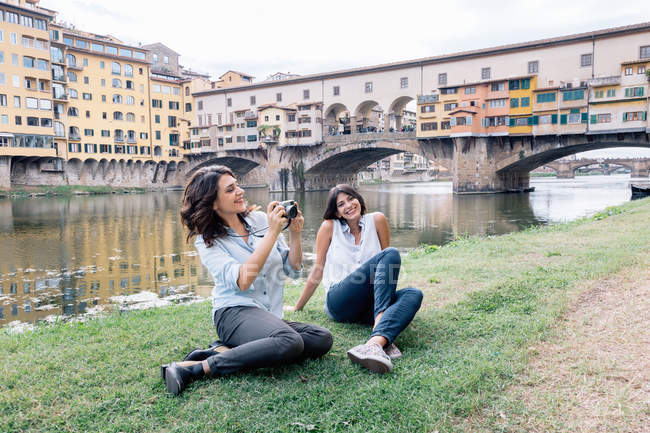 Casal de lésbicas sentado na margem do rio Arno em frente à Ponte Vecchio segurando câmera digital sorrindo, Florença, Toscana, Itália — Fotografia de Stock