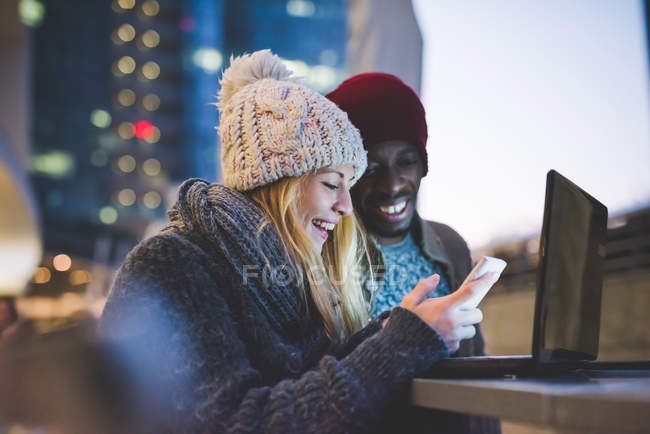 Пара с помощью смартфона и ноутбука на открытом воздухе в сумерках — стоковое фото