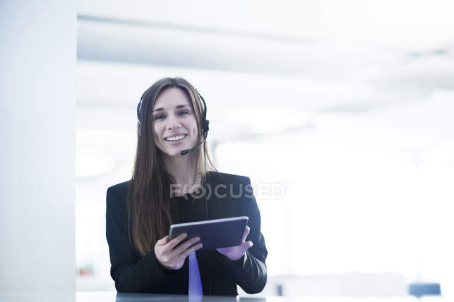 Молода жінка в телефонній гарнітурі тримає цифровий планшет, дивлячись на камеру посміхаючись — стокове фото