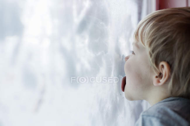 Garçon lécher à l'intérieur de la fenêtre couverte de neige en hiver — Photo de stock