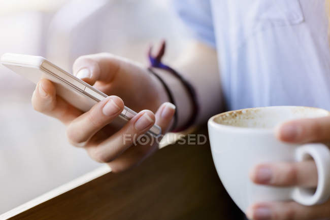 Cropped view of young womans hands holding coffee cup using smartphone — Stock Photo