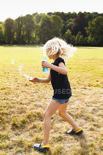 Chica ondeando varita de burbujas y haciendo burbujas en el parque - foto de stock