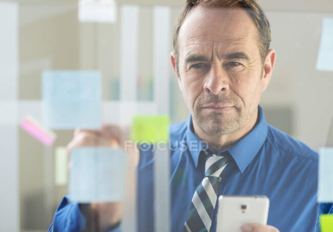 Empresário com notas de adesivo de smartphone na parede de vidro do escritório — Fotografia de Stock
