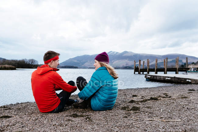 Pareja joven senderismo, sentado junto al borde de las aguas, Derwent Water, Keswick, Lake District, Cumbria, Reino Unido - foto de stock