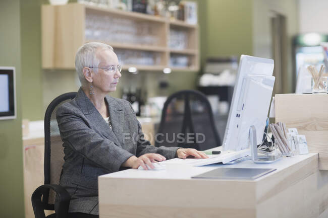 Recepcionista femenina senior usando computadora de escritorio en la recepción del hotel - foto de stock