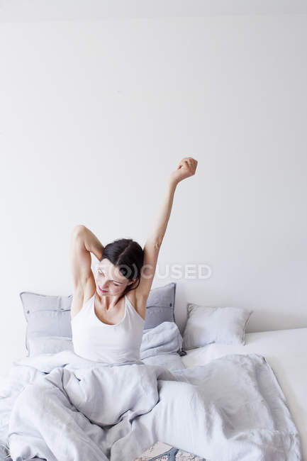 Mulher madura vestindo colete sentado na cama debaixo de braços de colcha levantada alongamento — Fotografia de Stock