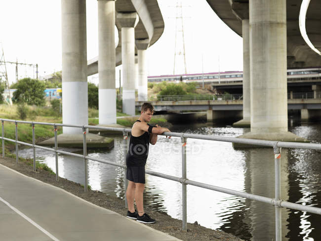 Молодий чоловік біля річки, спирається на перила, використовуючи MP3-плеєр, прикріплений до руки — стокове фото