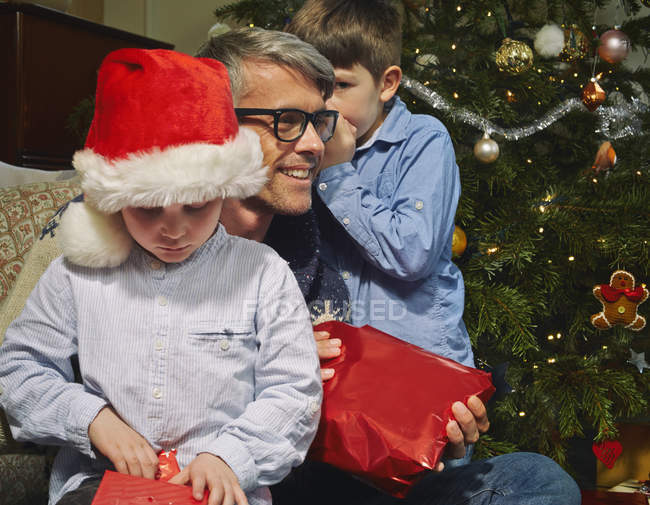 Junge flüstert Vater zu, während Bruder Weihnachtsgeschenke öffnet — Stockfoto
