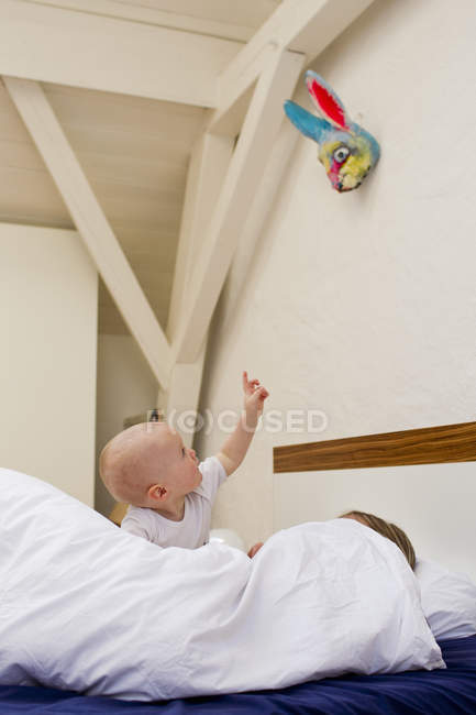Baby girl pointing from bed whilst mother under covers — Stock Photo