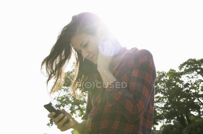 Mujer joven leyendo textos de teléfonos inteligentes contra el cielo soleado - foto de stock