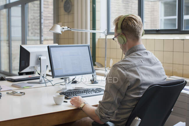 Visão traseira do homem ouvindo fones de ouvido enquanto trabalhava na mesa de escritório — Fotografia de Stock