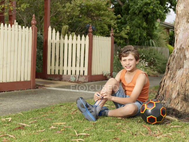 Retrato de menino com bola de futebol sentado na grama e amarrando atacadores — Fotografia de Stock
