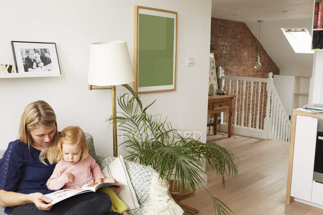 Woman and toddler daughter reading book on living room sofa — Stock Photo
