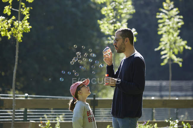 Hombre soplando burbujas como hija mira hacia arriba - foto de stock