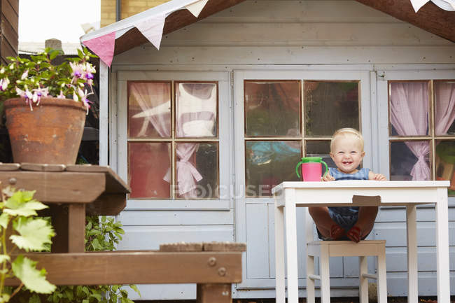 Baby-Mädchen sitzt am Tisch vor Spielhaus und blickt lächelnd in die Kamera — Stockfoto