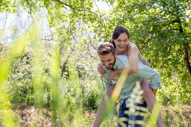 View through foliage of young man giving young woman piggyback — Stock Photo