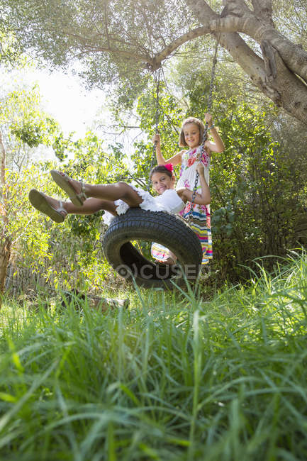 Two girls sitting and standing on tree swing in garden — Stock Photo