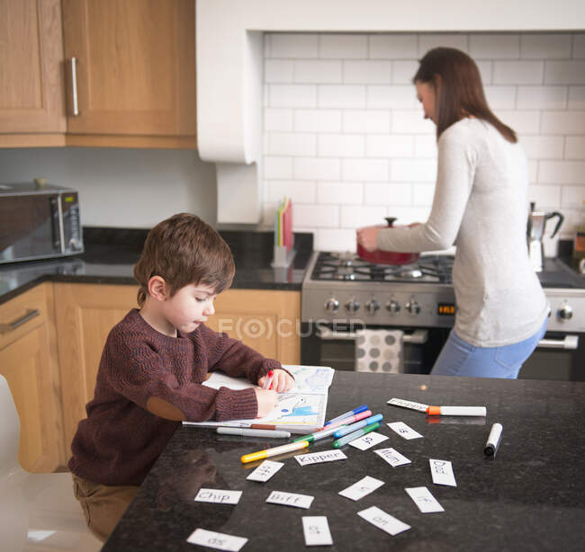 Niño para colorear en el libro en el mostrador de la cocina como madre prepara la cena - foto de stock