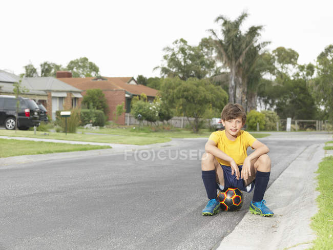 Portrait d'un garçon assis sur un ballon de football sur une route de banlieue — Photo de stock