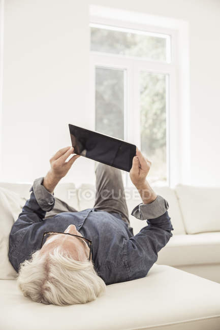 Homem sênior deitado no sofá, usando tablet digital, visão traseira — Fotografia de Stock