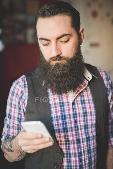 Hombre barbudo joven usando smartphone en la habitación - foto de stock