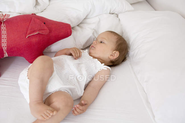 Baby girl lying on bed looking at soft toy — Stock Photo