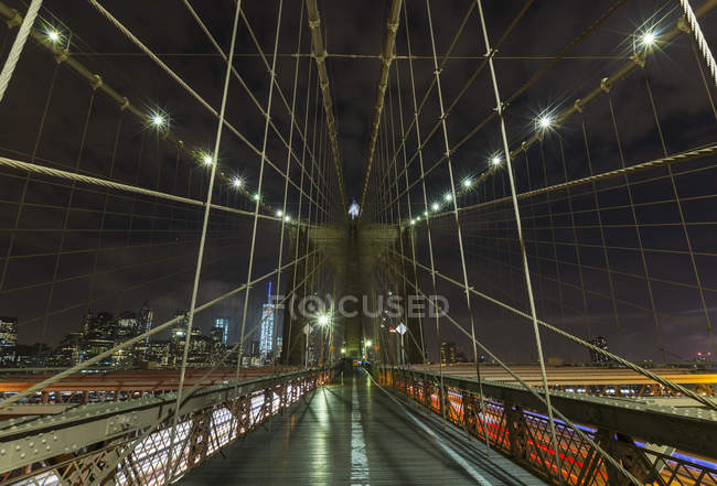 Brooklyn bridge walkway and distant Manhattan financial district skyline at night, New York, USA — Stock Photo