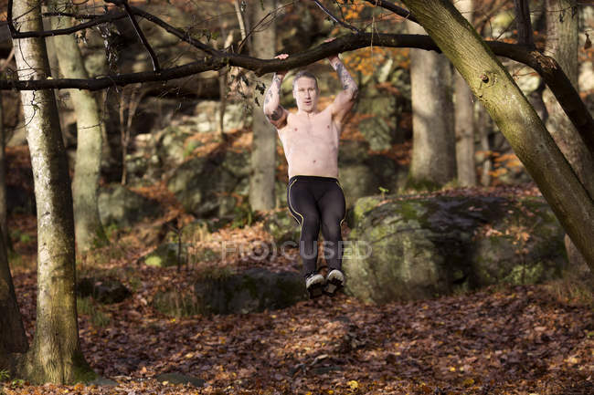 Comprimento total vista frontal do homem adulto médio tatuado na floresta fazendo puxar para cima na árvore — Fotografia de Stock