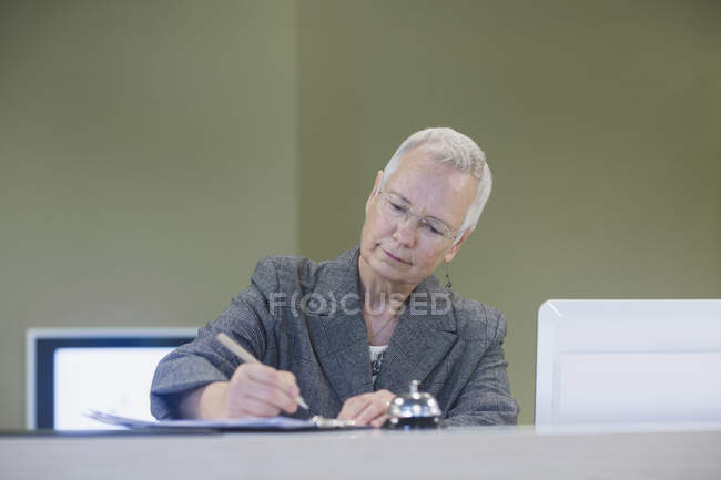 Senior female receptionist writing at hotel reception desk — Stock Photo