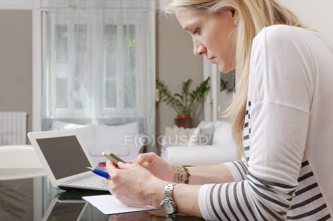 Молода жінка читає текстове повідомлення на смартфоні на домашньому столі — стокове фото