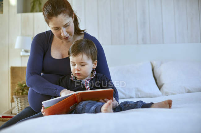 Mère enceinte et bébé garçon assis sur le lit lecture livre ensemble — Photo de stock