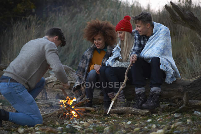 Four adult friends burning driftwood campfire on beach at dusk — Stock Photo