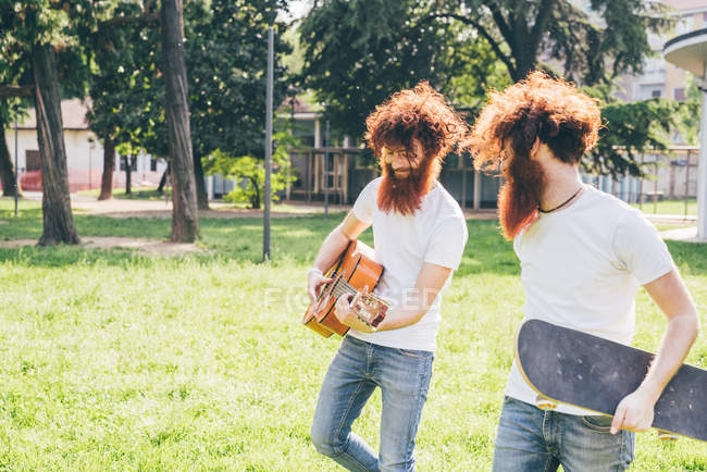 Молодые хипстеры-близнецы с рыжими волосами и бородами прогуливаются в парке, играя на гитаре — стоковое фото
