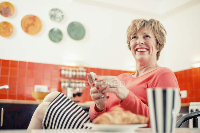 Зріла жінка тримає мобільний телефон на кухні, портрет — стокове фото