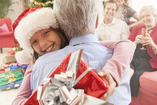 Neta segurando presente de Natal abraçando avô sorrindo — Fotografia de Stock