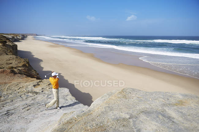 Golfer steht auf Klippe mit Blick auf Strand und nimmt Golfschwung — Stockfoto
