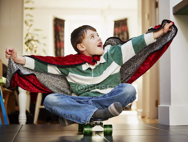 Jeune garçon assis sur une planche à roulettes portant une cape — Photo de stock