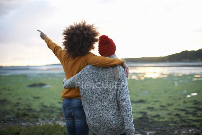 Visão traseira de duas jovens mulheres por mar apontando ao pôr do sol — Fotografia de Stock