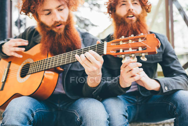 Junge männliche Hipster-Zwillinge mit roten Bärten sitzen und spielen Gitarre — Stockfoto