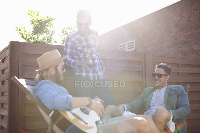 Tres amigos masculinos charlando y tocando la guitarra en la fiesta en la azotea - foto de stock