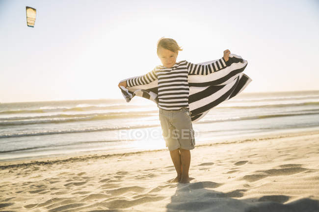 Повнометражний вид спереду хлопчика на пляжі в смугастій футболці, що тримає рушник, дивлячись на камеру — стокове фото