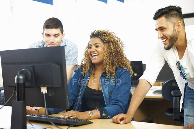 Three student friends laughing and looking at personal computer in classroom — Stock Photo