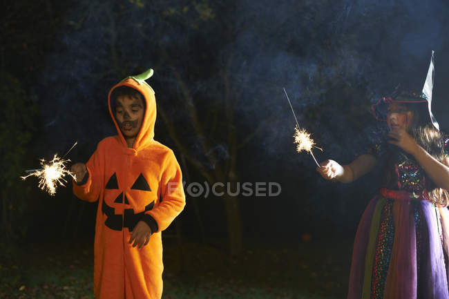 Hermano y hermana vistiendo disfraces de Halloween sosteniendo bengalas en el jardín por la noche - foto de stock