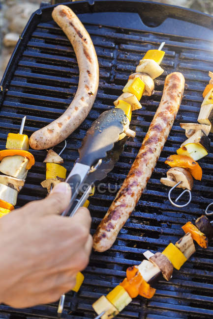 Persona che utilizza pinze per trasformare salsicce e spiedini su griglia barbecue — Foto stock